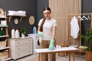 Photo of Beautiful woman with spray bottle and microfiber cloth cleaning white table in laundry room