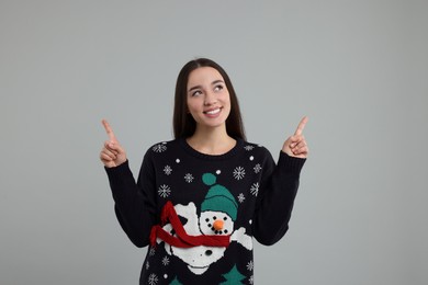 Happy young woman in Christmas sweater pointing at something on grey background