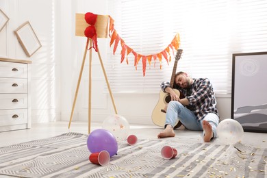 Young man with guitar sleeping near window in messy room after party