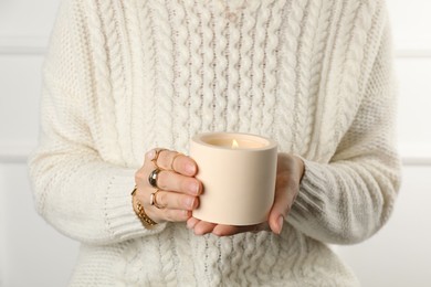 Woman with stylish jewelry holding burning soy candle in ceramic holder on light background, closeup