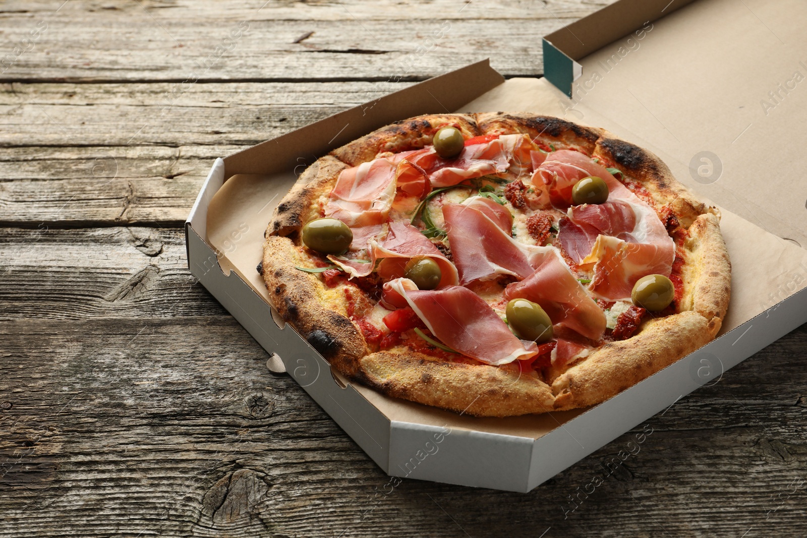 Photo of Tasty pizza with cured ham, olives and sun-dried tomato in cardboard box on wooden table, space for text