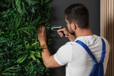 Photo of Man with screwdriver installing green artificial plant panel on grey wall in room