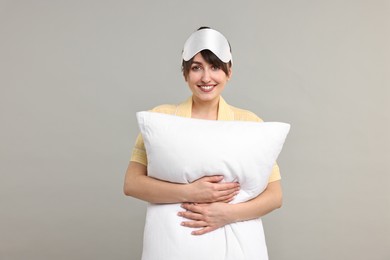 Photo of Happy woman in pyjama and sleep mask holding pillow on grey background