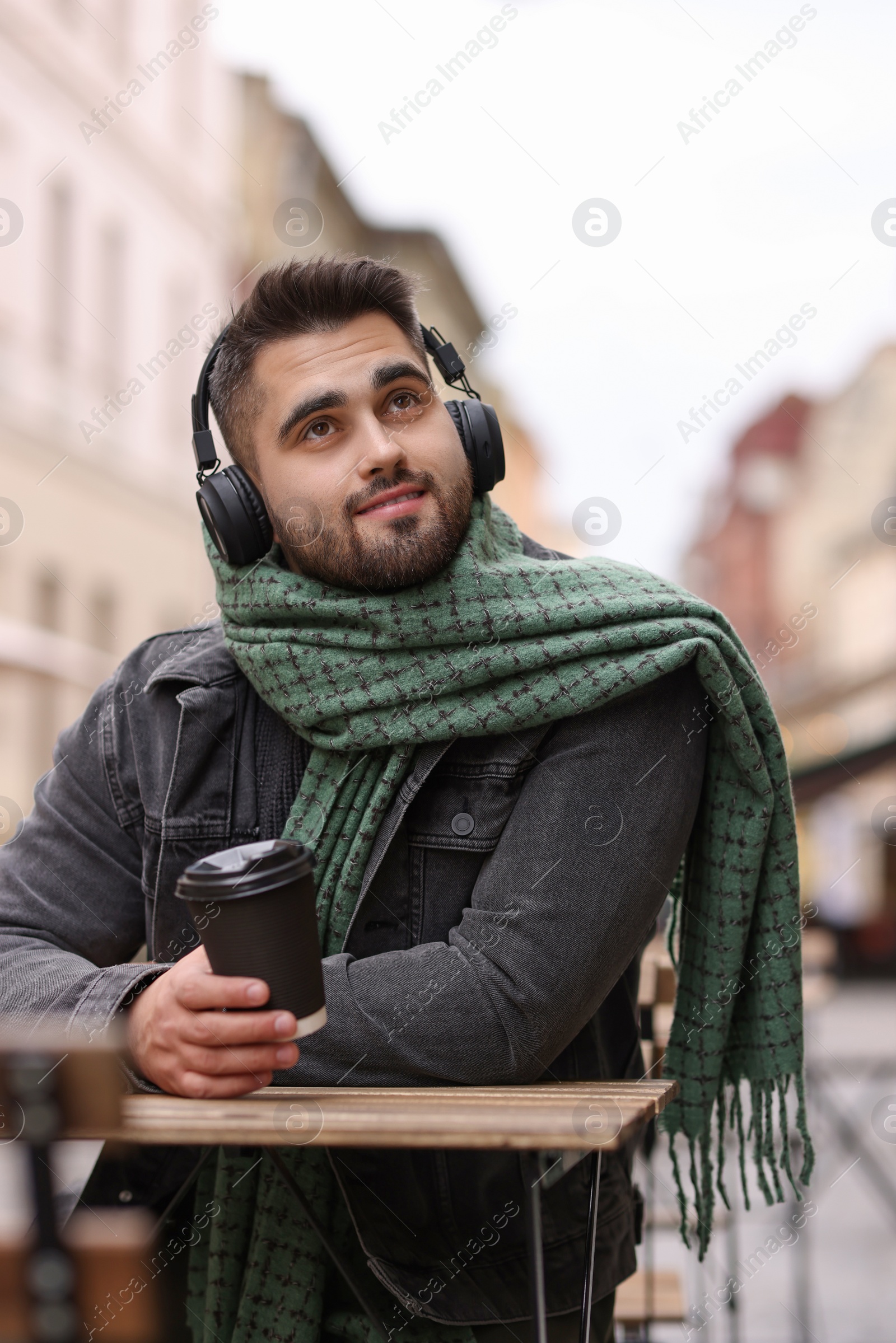 Photo of Smiling man in warm scarf with paper cup listening to music in outdoor cafe