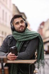 Smiling man in warm scarf with paper cup listening to music in outdoor cafe