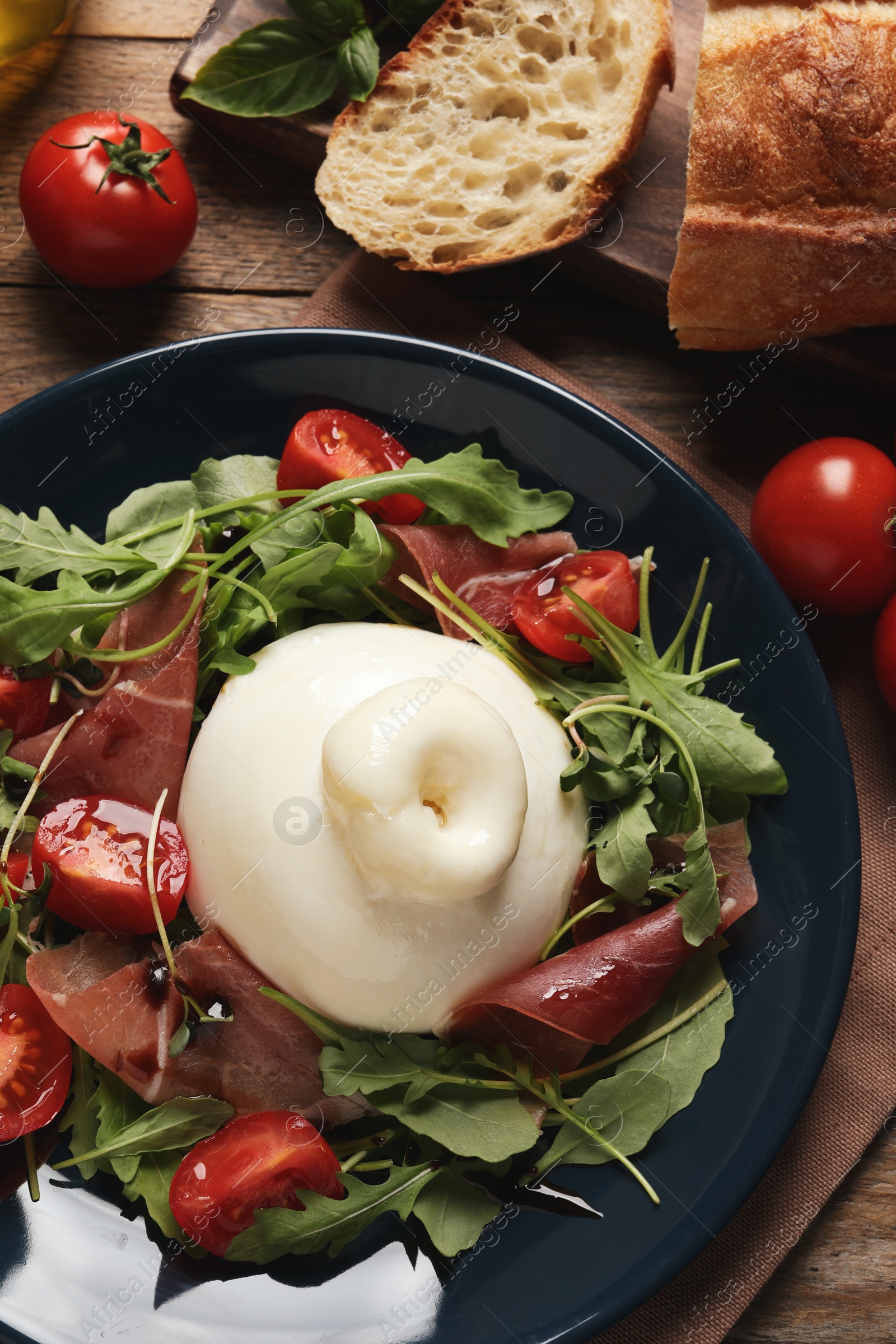 Photo of Delicious burrata salad with tomatoes, prosciutto and arugula on wooden table, flat lay