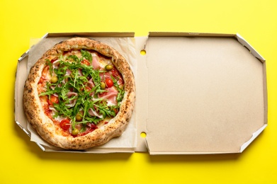 Photo of Tasty pizza with meat and arugula in cardboard box on yellow background, top view