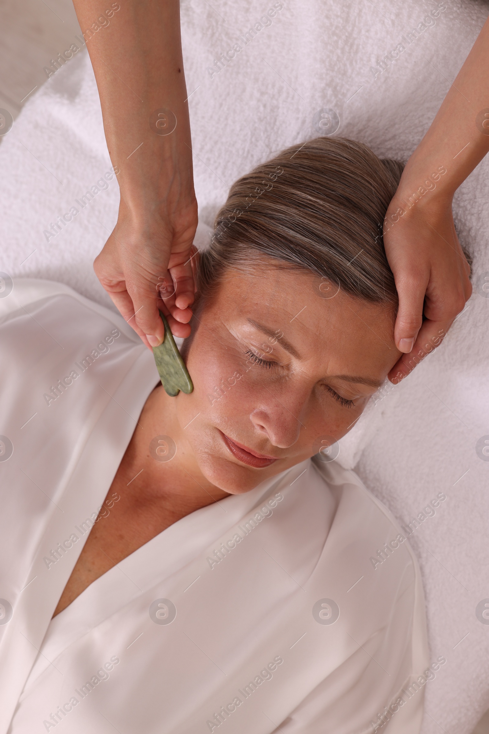 Photo of Woman receiving facial massage with jade gua sha tool in beauty salon, above view