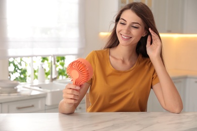 Photo of Woman enjoying air flow from portable fan at table in kitchen. Summer heat