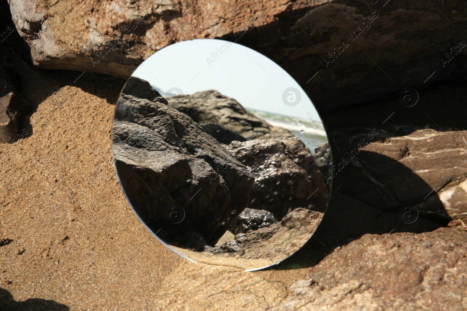 Photo of Round mirror reflecting beach on sand near stones outdoors