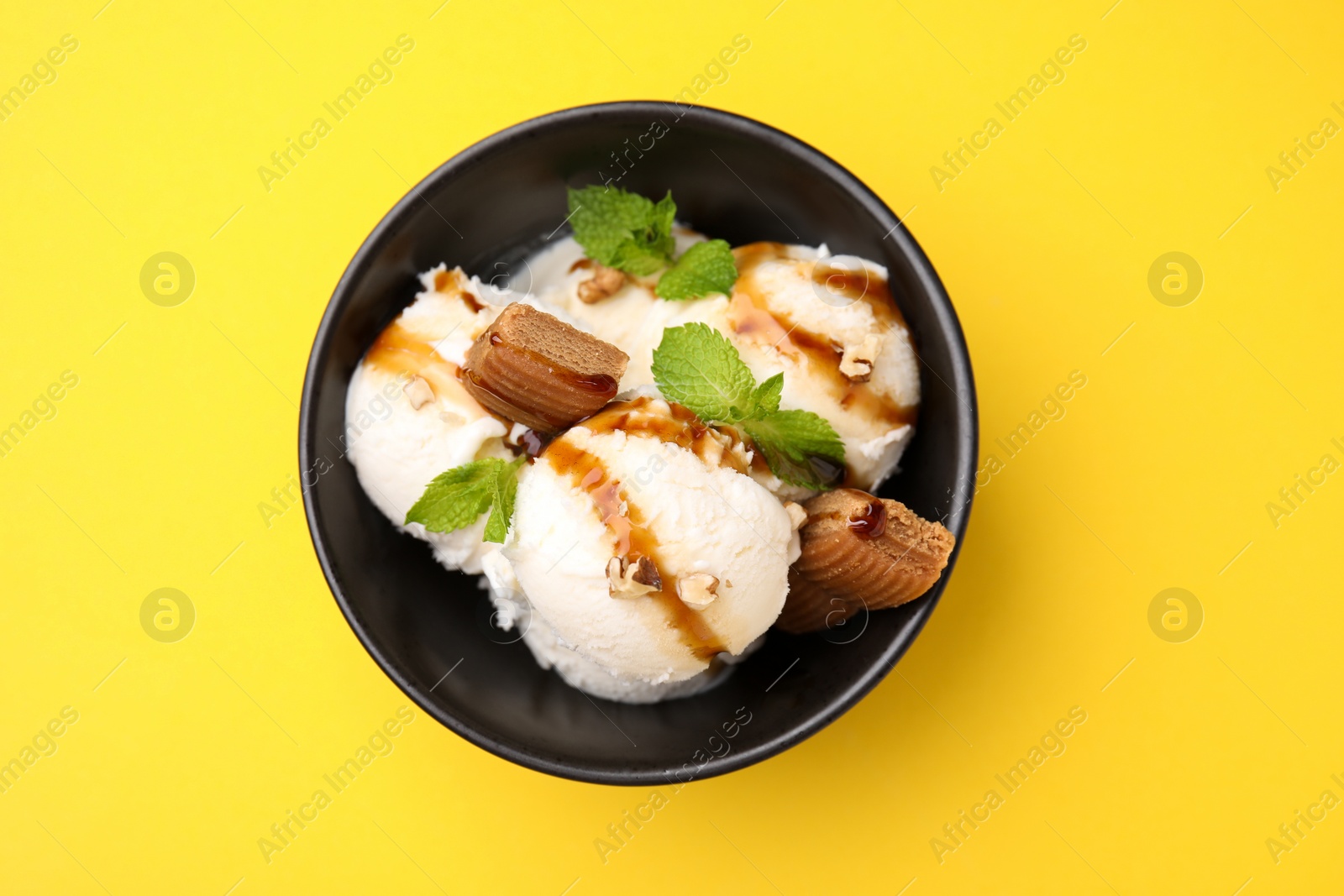 Photo of Tasty ice cream with caramel sauce, mint, candies and nuts in bowl on yellow background, top view