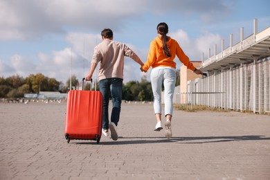 Being late. Couple with red suitcase running outdoors, back view