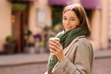 Beautiful woman in warm scarf with paper cup of coffee on city street, space for text