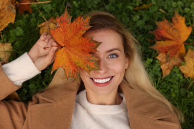 Smiling woman lying on grass and covering eye with autumn leaf, top view