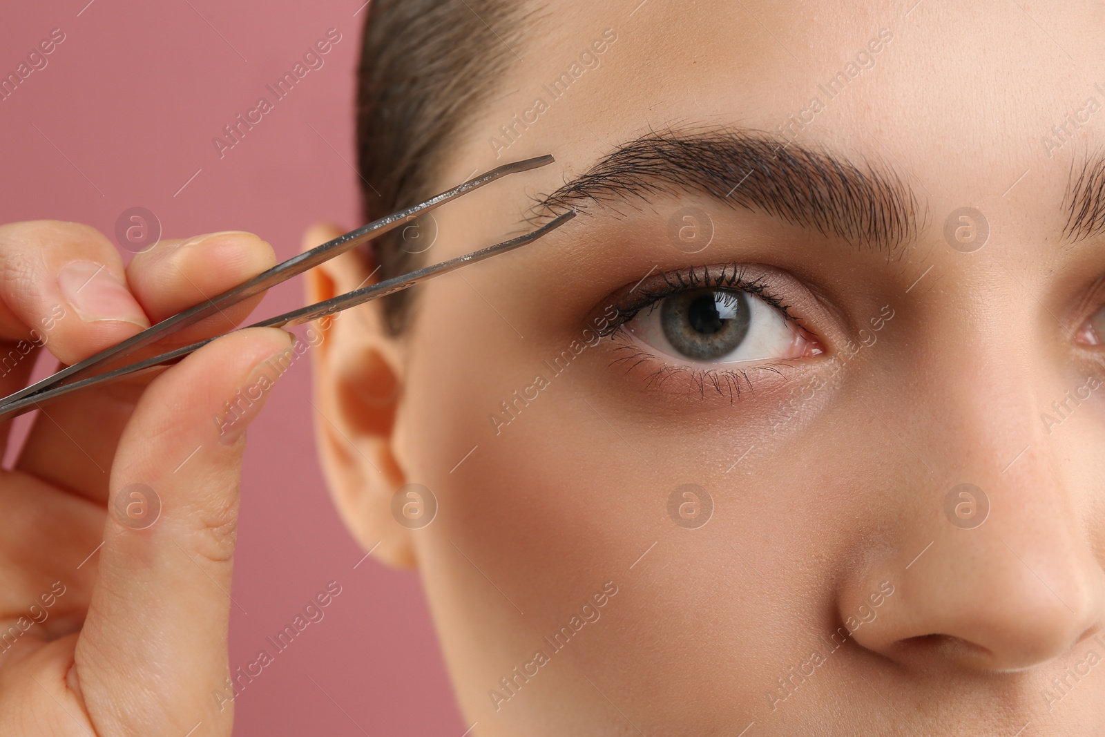 Photo of Eyebrow correction. Young woman with tweezers on pink background, closeup