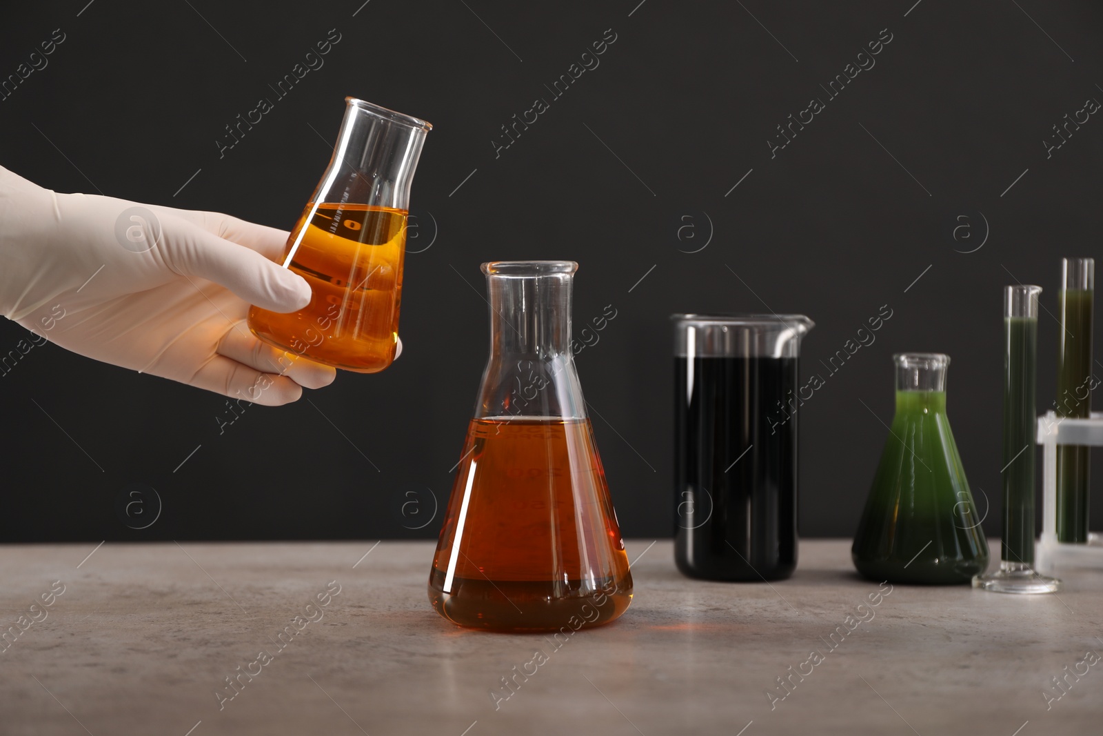 Photo of Woman holding flask with yellow crude oil over grey table against dark background, closeup