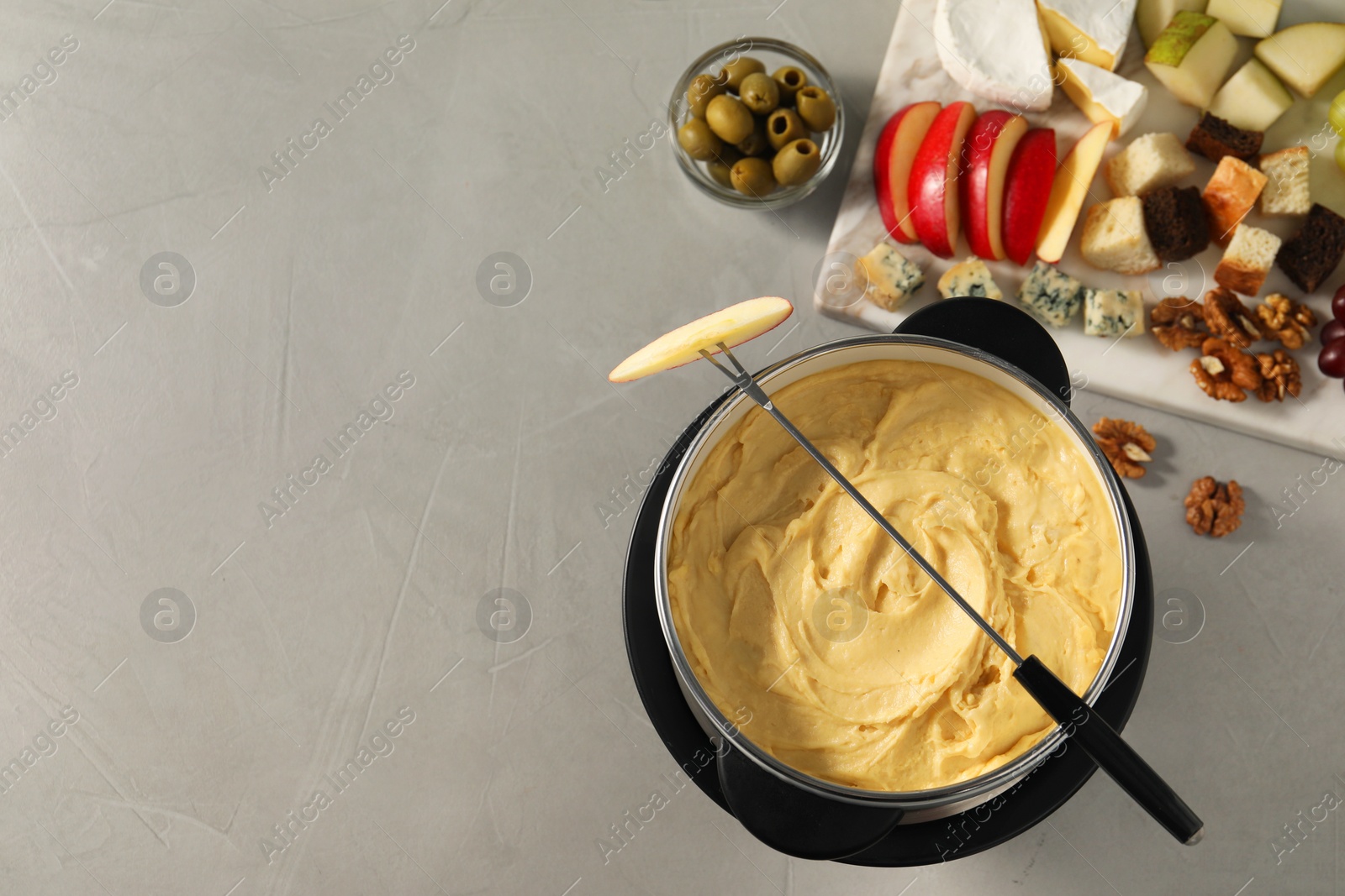 Photo of Fork with piece of apple, melted cheese in fondue pot and snacks on grey table, flat lay. Space for text