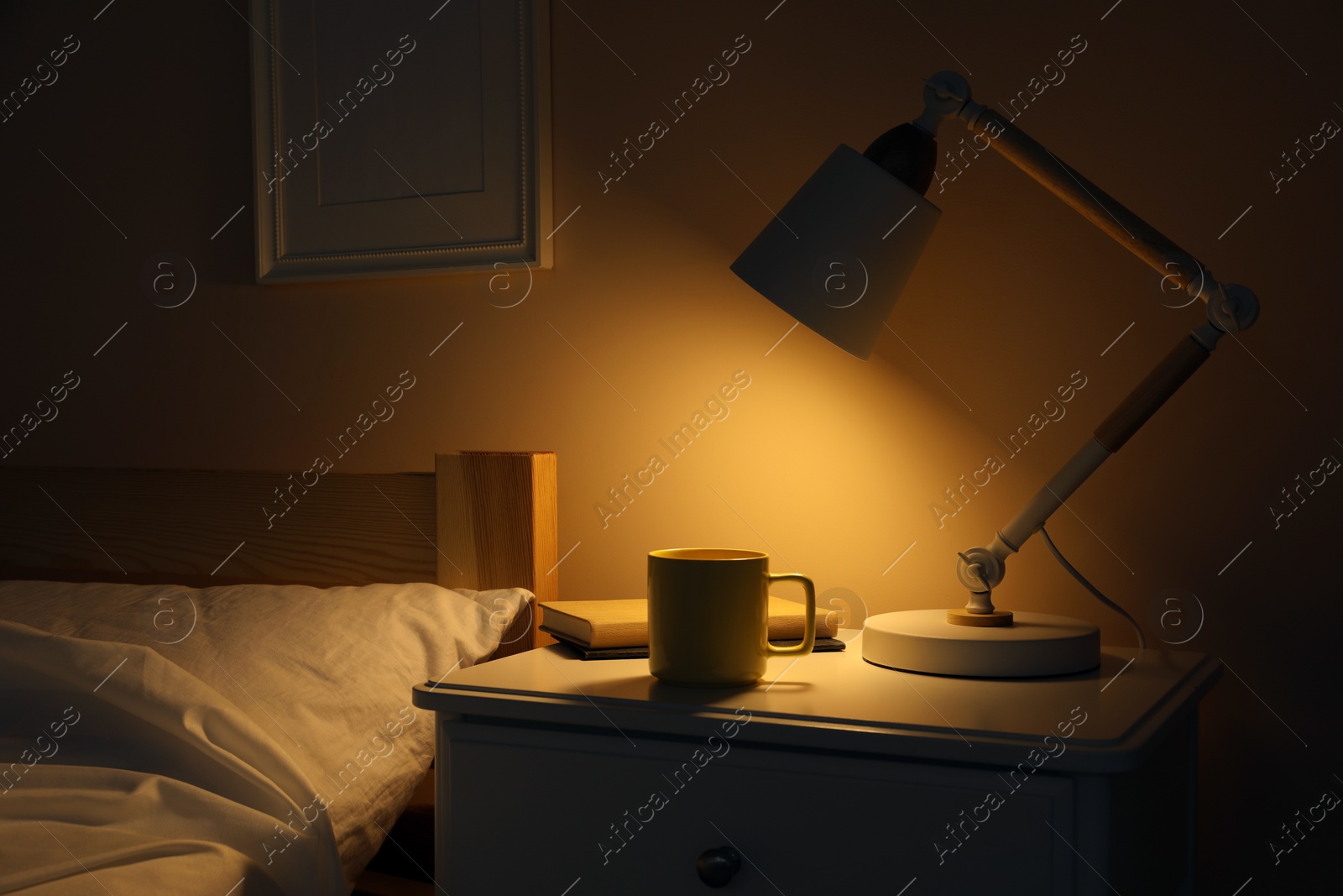 Photo of Stylish modern desk lamp, books and cup of drink on white nightstand in dark bedroom