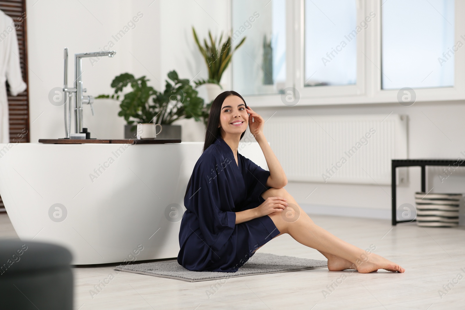 Photo of Beautiful happy woman wearing stylish bathrobe on floor near tub in bathroom