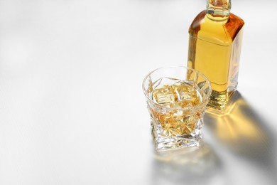 Whiskey with ice cubes in glass and bottle on white table, space for text