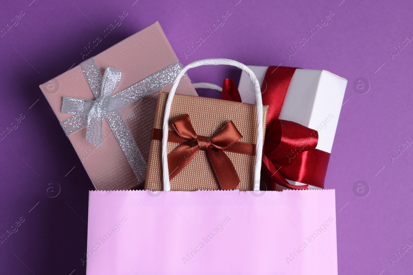Photo of Pink paper shopping bag full of gift boxes on purple background, flat lay