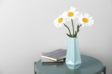 Beautiful tender chamomile flowers in vase and notebooks on table against light background, space for text