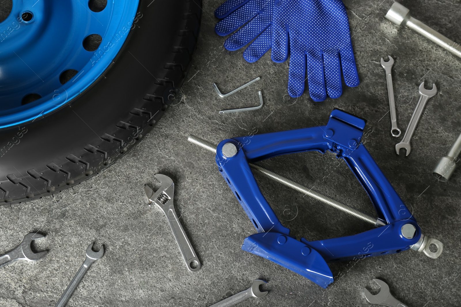Photo of Car wheel, scissor jack, gloves and different tools on grey stone surface, flat lay