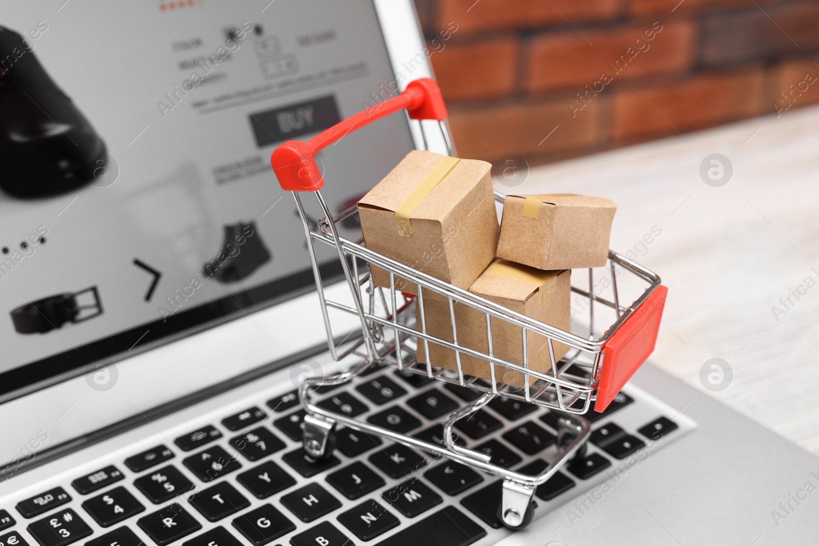 Photo of Internet store. Small cardboard boxes, shopping cart and laptop on light wooden table, closeup