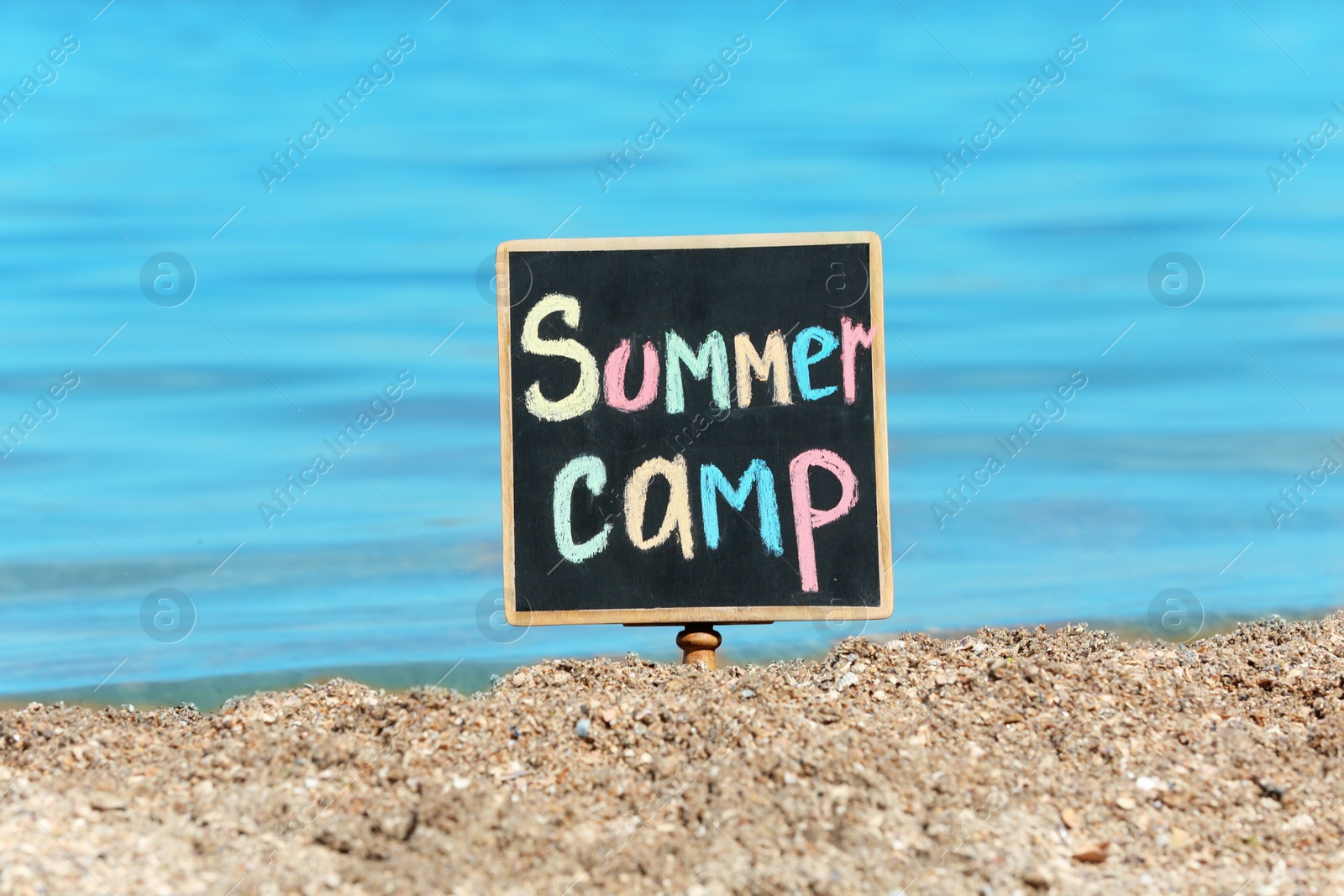 Photo of Little blackboard with text SUMMER CAMP on sand near sea