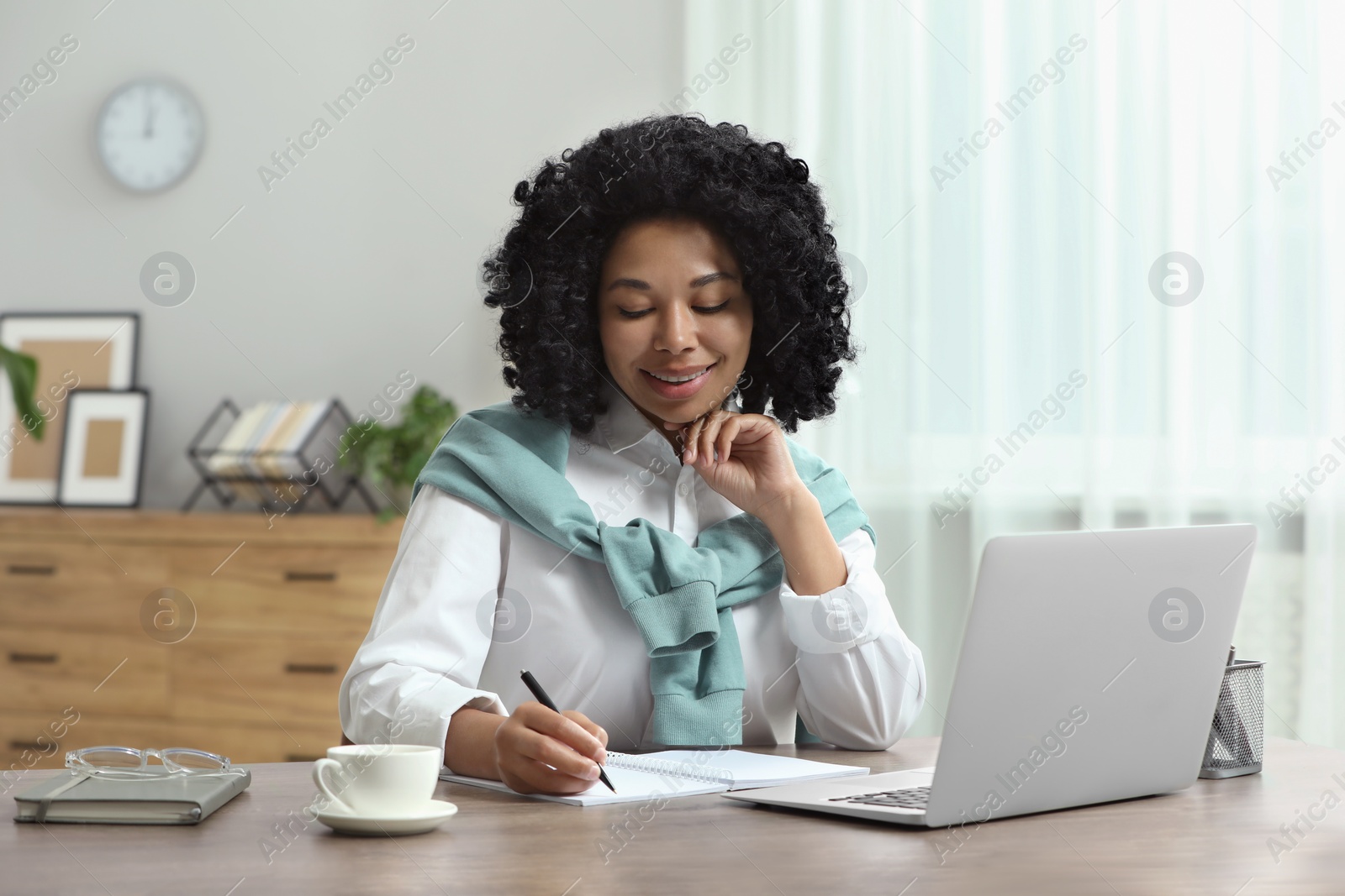 Photo of Happy young woman writing notes while using laptop at wooden desk indoors