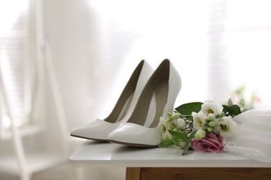 Photo of Pair of white high heel shoes, flowers and wedding dress on table indoors