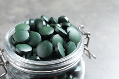 Glass jar with green spirulina pills on grey table, closeup