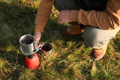 Man making hot drink with portable gas burner on green grass, closeup. Camping season