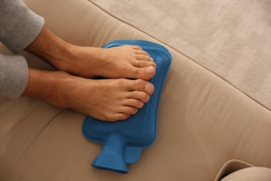 Photo of Man warming feet with hot water bottle on sofa, closeup