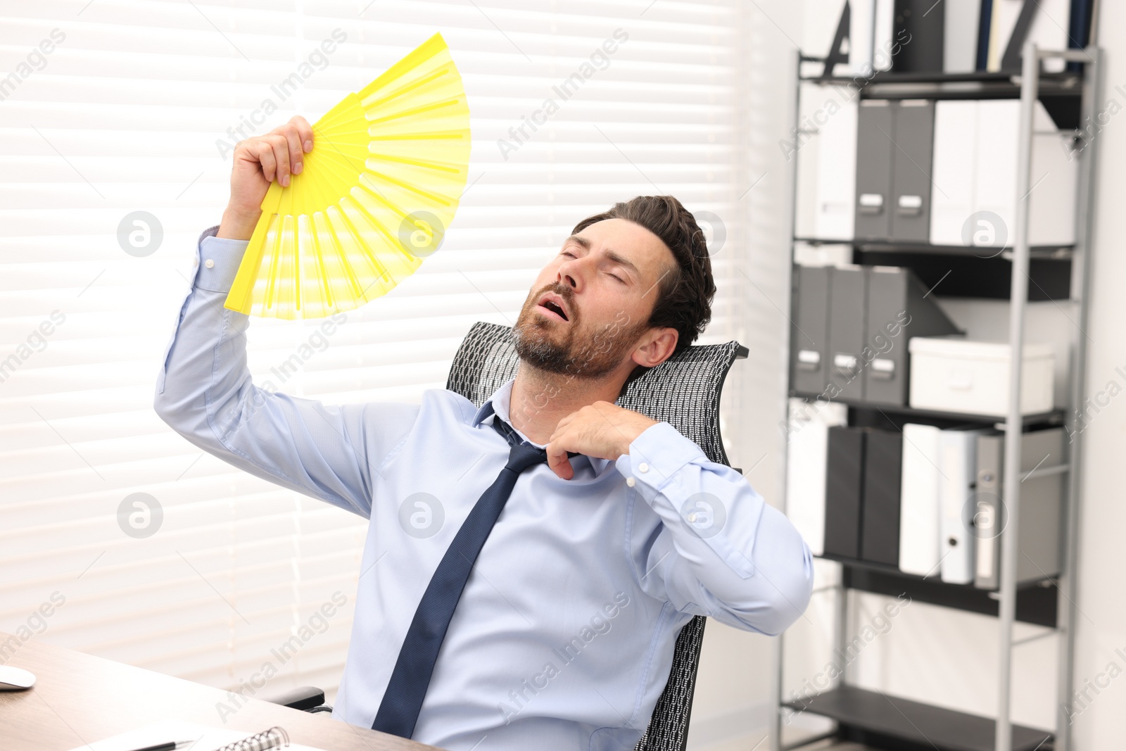 Photo of Bearded businessman waving yellow hand fan to cool himself in office