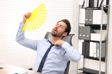 Bearded businessman waving yellow hand fan to cool himself in office