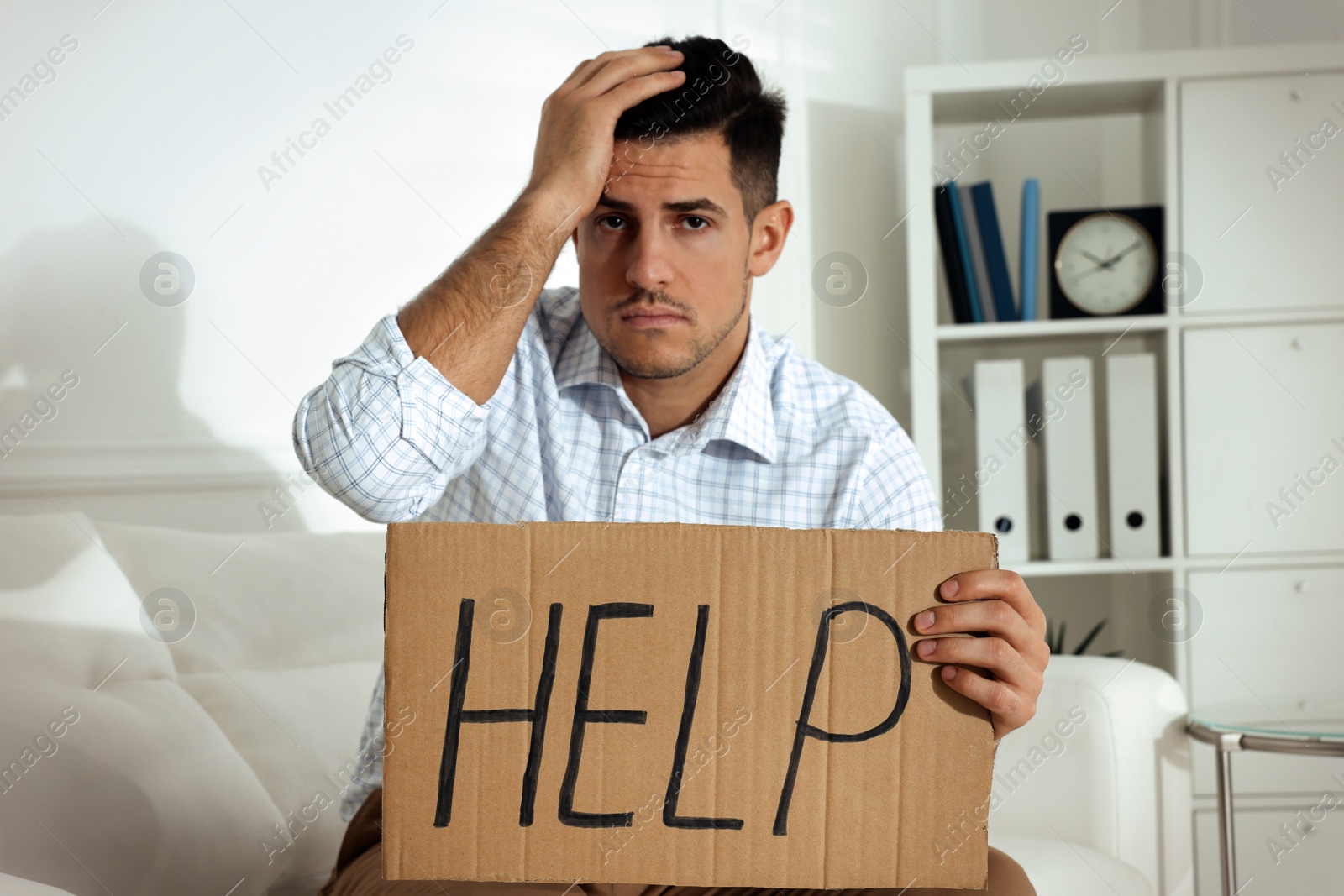 Photo of Unhappy man with HELP sign on sofa indoors