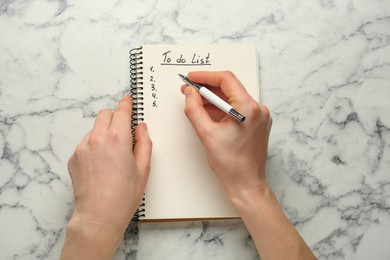 Woman filling To Do list in notepad at white marble table, top view