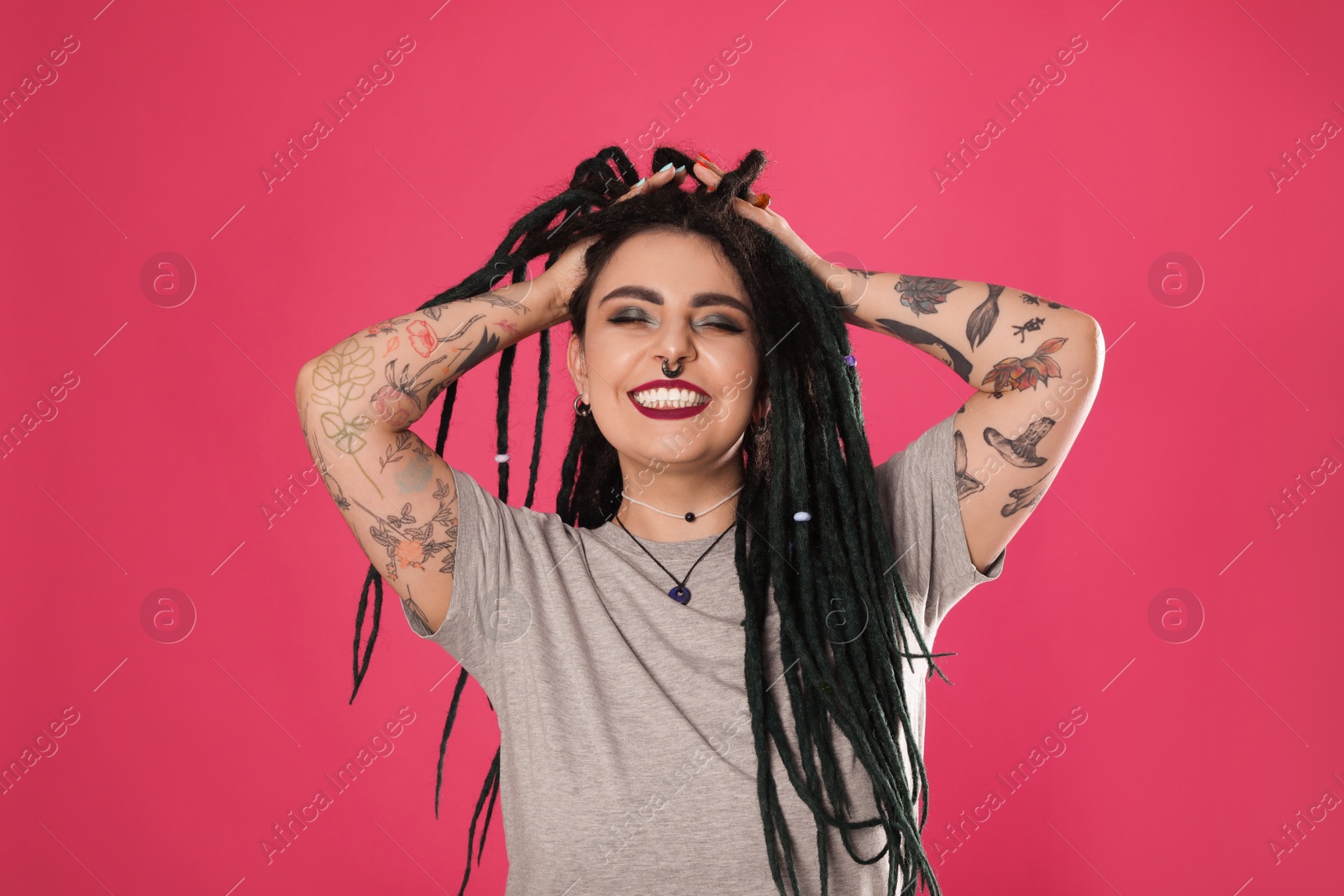 Photo of Beautiful young woman with tattoos on arms, nose piercing and dreadlocks against pink background