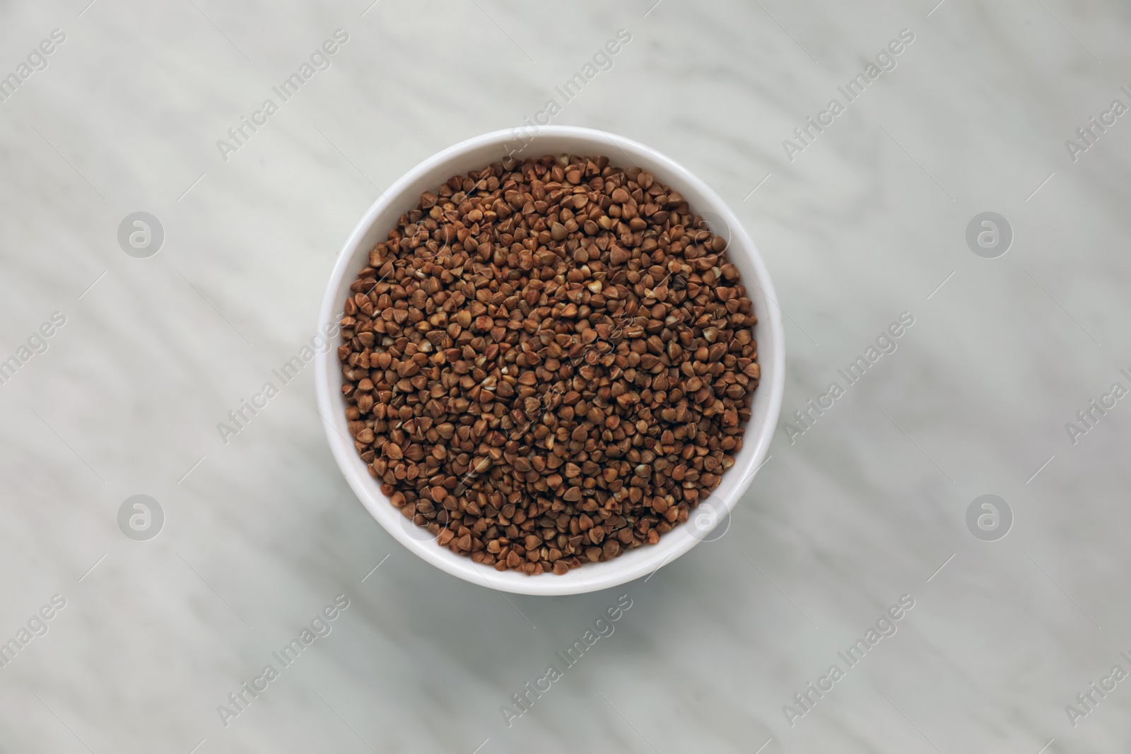 Photo of Bowl with uncooked buckwheat porridge on white marble table, top view