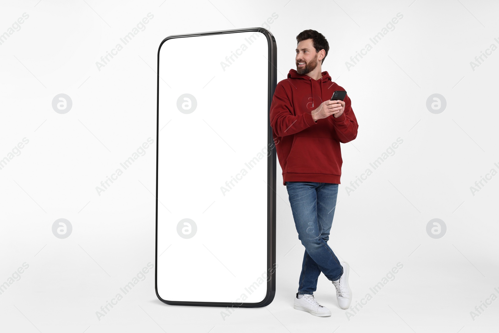 Image of Man with mobile phone standing near huge device with empty screen on grey background. Mockup for design