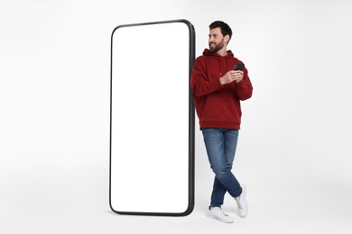 Man with mobile phone standing near huge device with empty screen on grey background. Mockup for design