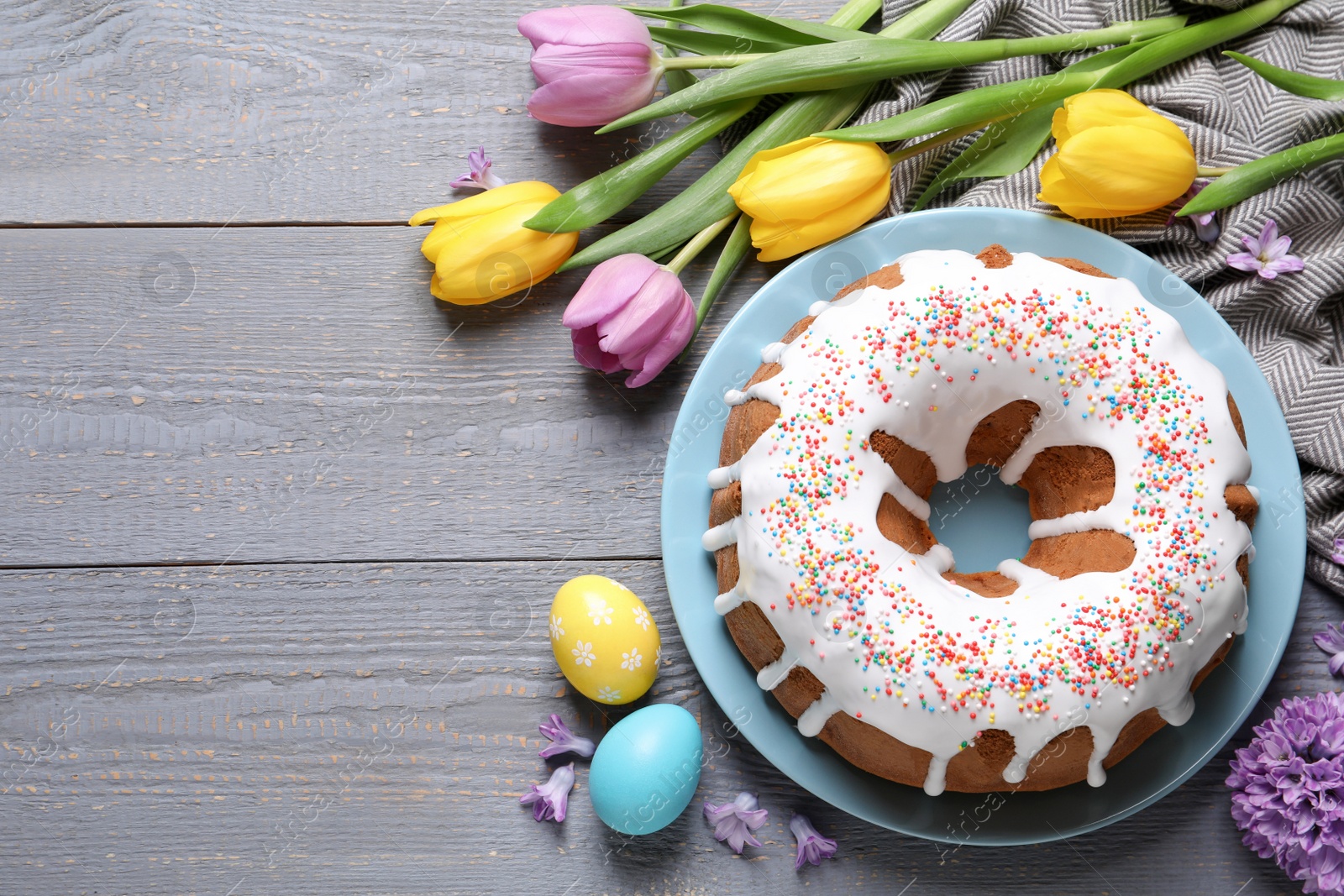 Photo of Glazed Easter cake with sprinkles, painted eggs and flowers on grey wooden table, flat lay. Space for text