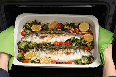 Woman taking baking dish with delicious fish and vegetables from oven, top view