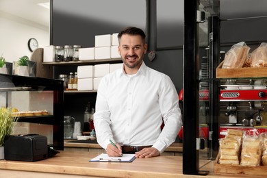 Photo of Business owner in his cafe. Man writing notes on clipboard near showcases with pastries