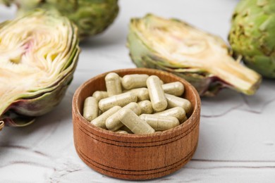 Bowl with pills and fresh artichokes on white textured table, closeup