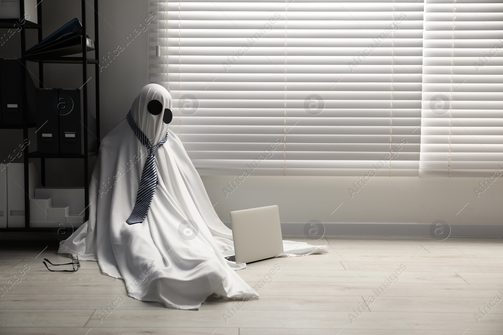 Photo of Overworked ghost. Man in white sheet with laptop on floor in office, space for text