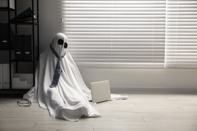 Overworked ghost. Man in white sheet with laptop on floor in office, space for text