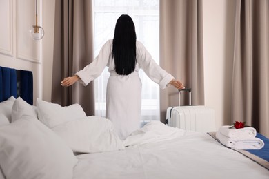 Photo of Young woman wearing bathrobe near window in hotel room, back view