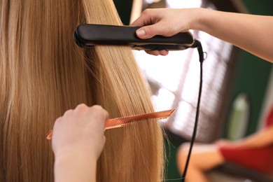 Stylist straightening woman's hair with flat iron in salon, closeup